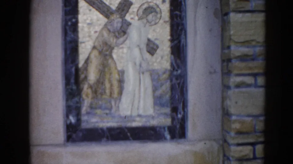 Final 8mm frame from 1962 Illinois church: mosaic of Jesus and companion under a cross in gold tile panel, dark marble border, stone wall and pilaster.