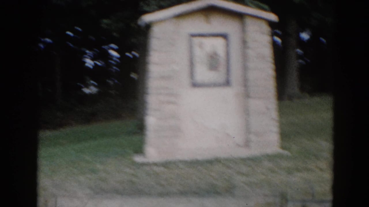 Small stone outdoor shrine on a grassy lawn with dark tree foliage behind, seen from a distance.