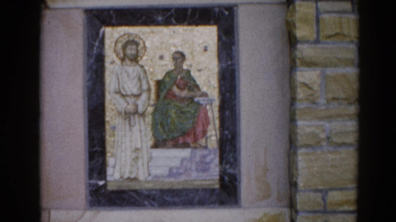 Close-up of a religious mosaic in a dark frame — a standing haloed figure and a seated figure in green-red robes on a gold ground.