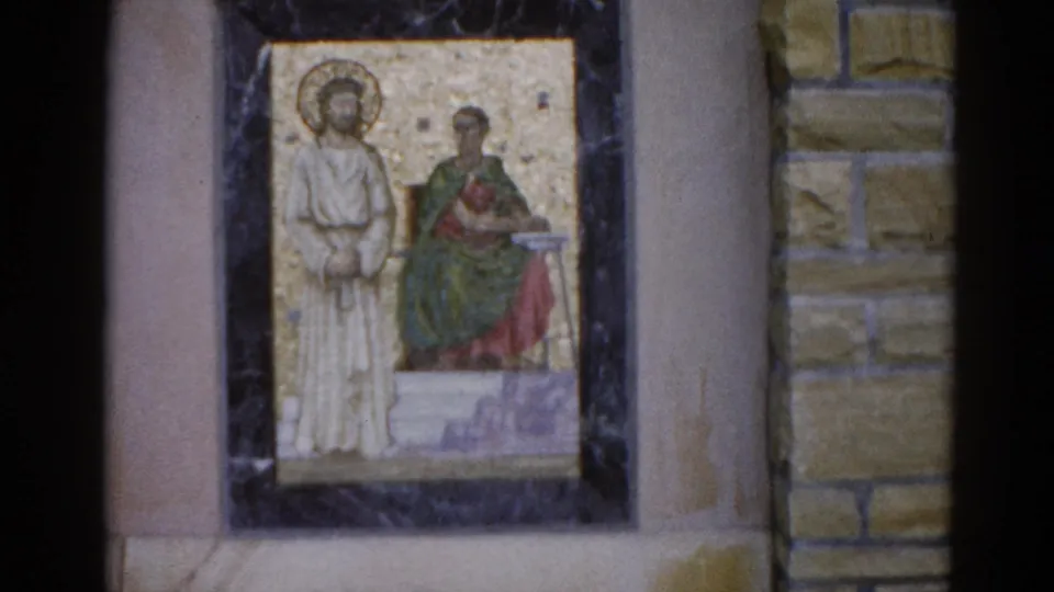 Religious mosaic icon in dark frame flanked by tan stone masonry blocks, two haloed figures, Illinois circa 1962; 8mm close-up with visible film wear.