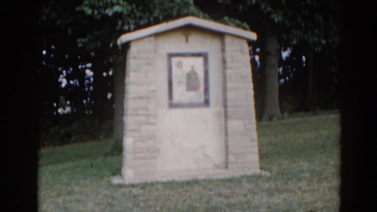 Small stone outdoor religious shrine with peaked roof and a framed devotional image showing a robed figure, set on a green lawn with trees, Illinois circa 1962.