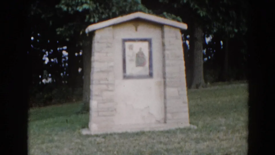 Small stone outdoor religious shrine with peaked roof and a framed devotional image showing a robed figure, set on a green lawn with trees, Illinois circa 1962.