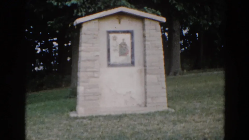 Stone outdoor religious shrine centered on a green lawn, framed devotional image visible on its face, trees in background, Illinois circa 1962, 8mm film.