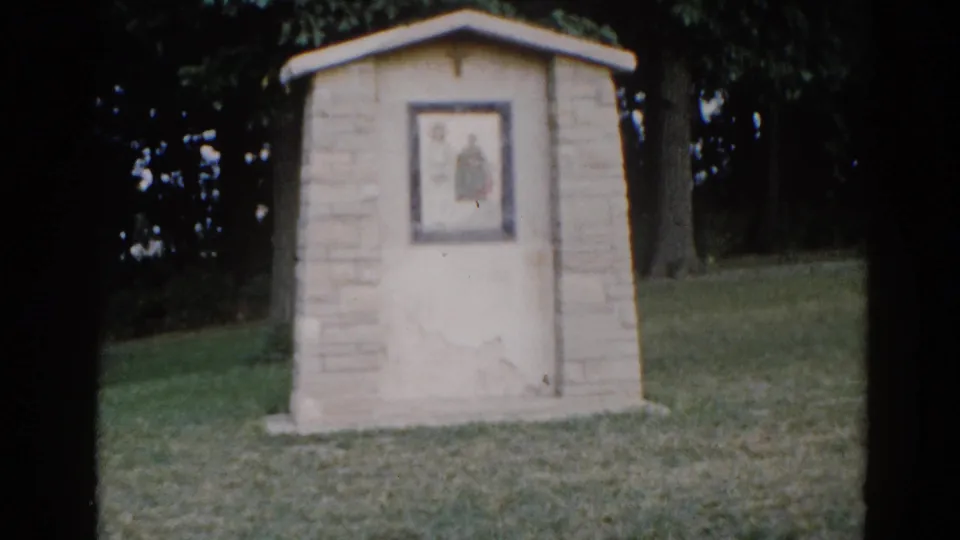 Stone outdoor religious shrine with framed devotional panel, green lawn and deciduous trees visible behind, Illinois circa 1962, 8mm film.