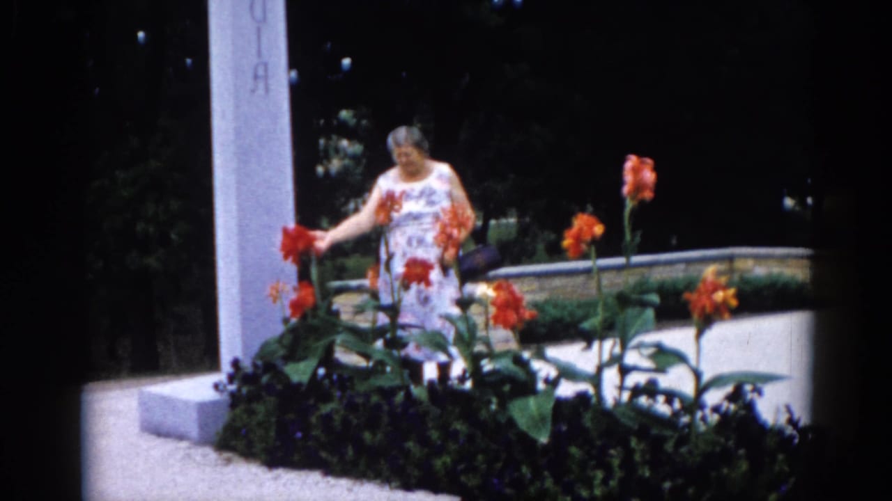 Older woman in a light floral dress touching tall orange-red flowers beside a raised stone planter and an inscribed white pillar — Illinois garden, c. 1962.
