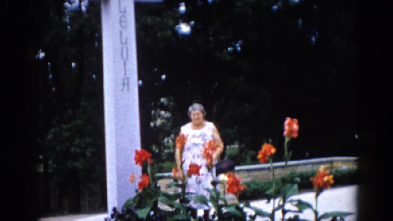 Older woman in floral dress posing among tall orange blooms with an inscribed white stone pillar at right — Illinois garden, c. 1962, 8mm.
