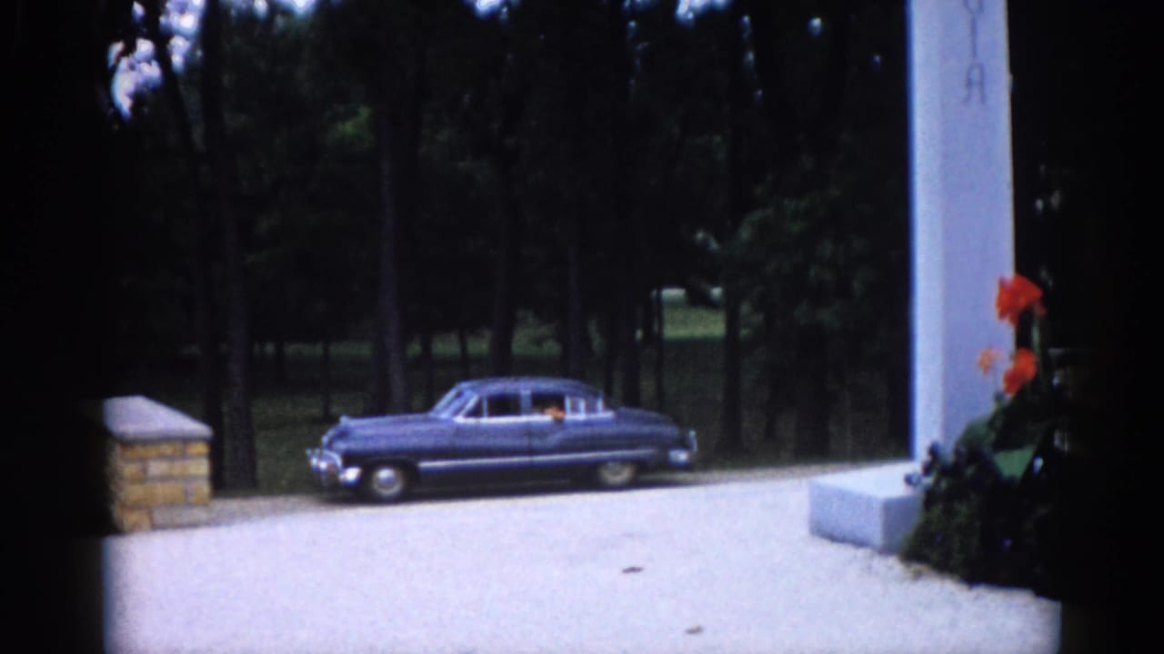 Gravel cemetery drive with a dark blue 1960s-era sedan parked beside a tall white pillar and orange canna lily bed, dense trees behind.
