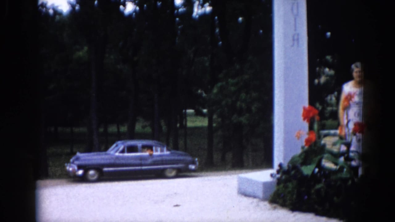 Elderly woman in white patterned dress partially visible at right beside tall white pillar; dark blue sedan and orange canna lilies in frame.
