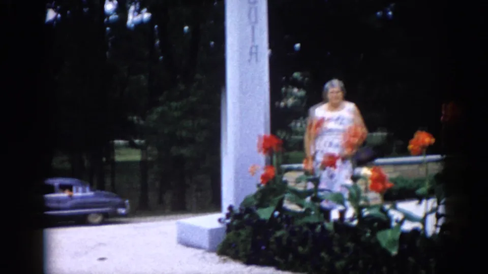 Early 1960s Illinois cemetery: older woman in white floral dress standing among vibrant orange canna lilies at a tall white pillar, sedan partially visible at left, 8mm grain.