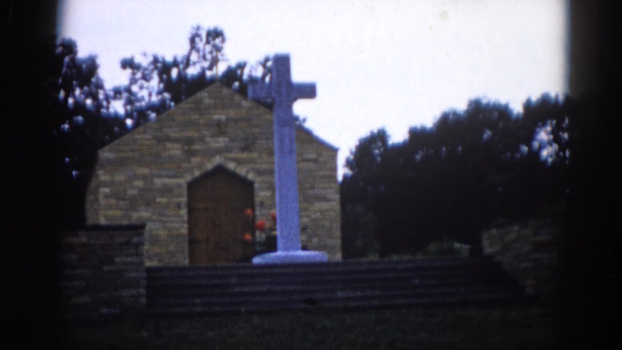 Stone brick chapel with a tall white cross on a pedestal in front, pointed arched wooden door visible, dark trees behind, 8mm film vignetting.