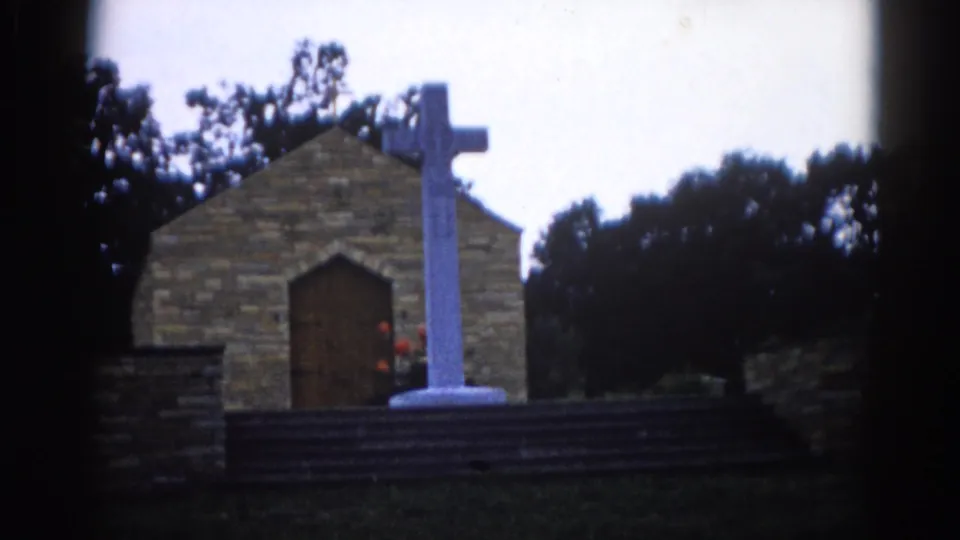 Exterior of a small Illinois brick chapel circa 1962, white cross on a stone base stands before the arched wooden door, overcast sky and trees behind.