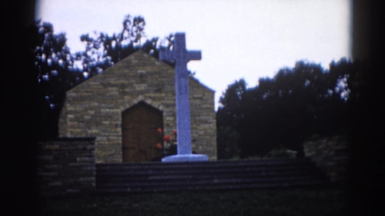 Same static exterior view of brick chapel and white cross, arched wooden door and red flowers at base visible, leafy tree canopy behind.