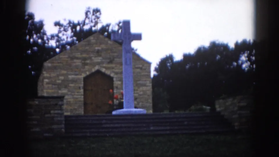 Small Illinois chapel exterior filmed on 8mm circa 1962, white cross on pedestal before a pointed arched door, muted tones and edge vignetting.