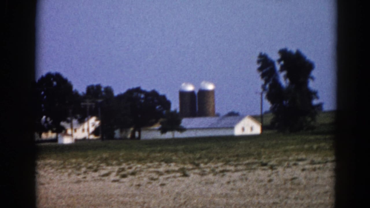 Rural farmstead with white buildings, corrugated metal barn, and two grain silos on a gentle hill under a blue-grey sky.