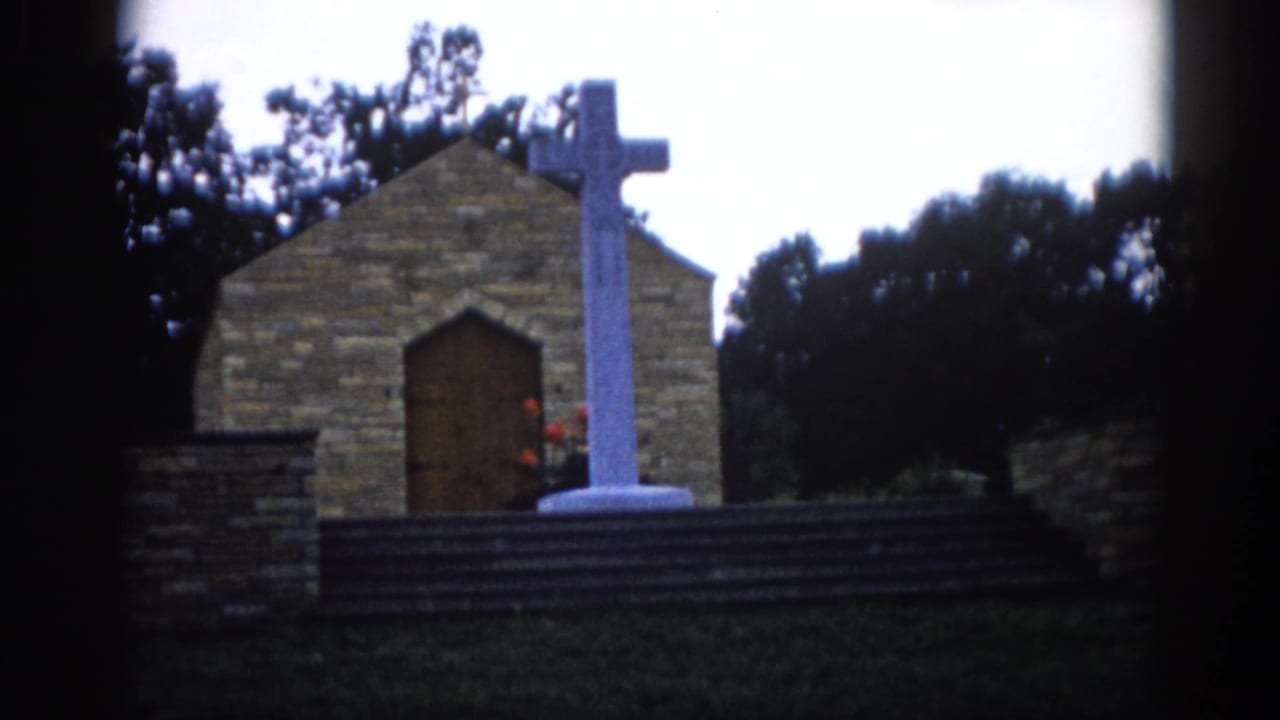 Stone chapel with arched wooden door and large white Latin cross on a stepped base, surrounded by trees.