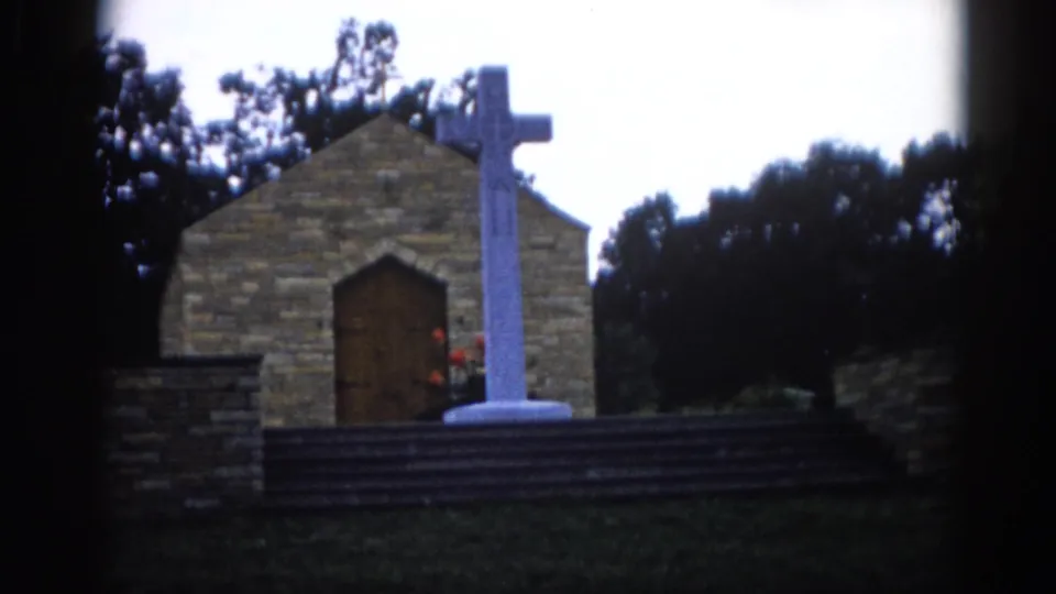 8mm home movie frame, c. 1962, Illinois — rough-cut stone chapel exterior with large white cross on stepped base and arched wooden door; red flowers visible at cross base.