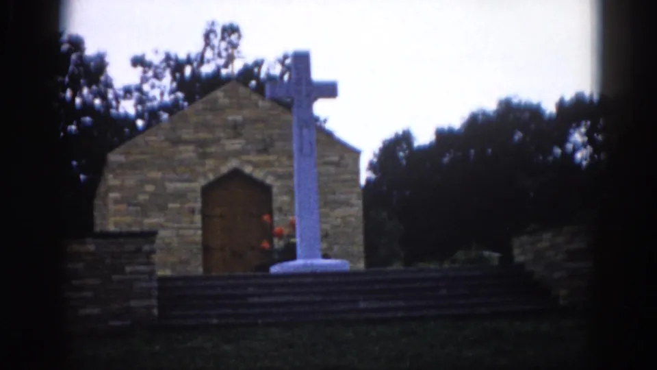 8mm home movie frame, c. 1962, Illinois — wider shot of same stone chapel and white cross with green lawn in foreground and surrounding trees.