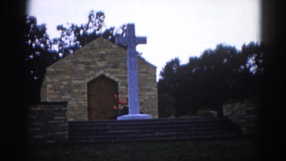 8mm home movie frame, c. 1962, Illinois — stone chapel exterior with white cross; composition nearly identical to prior frame with slight camera drift.