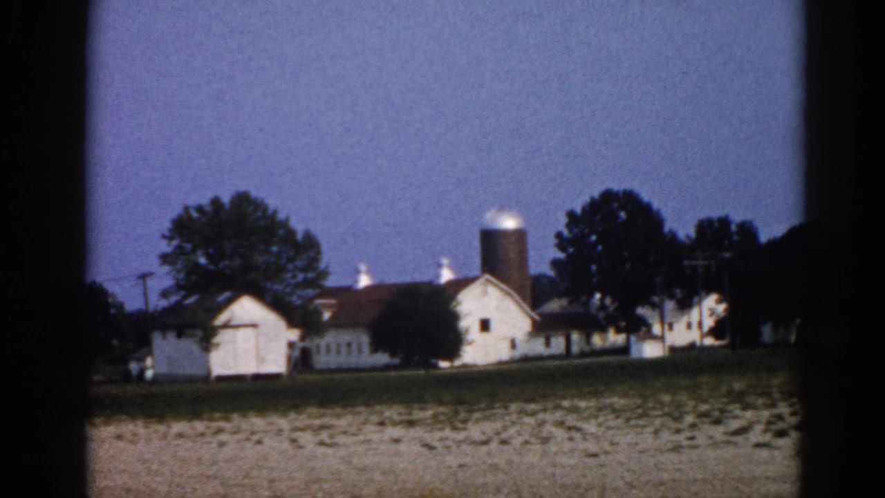 White barn and grain silos at dusk, viewed through a dark-bordered film frame, with a dry foreground field and blue twilight sky.
