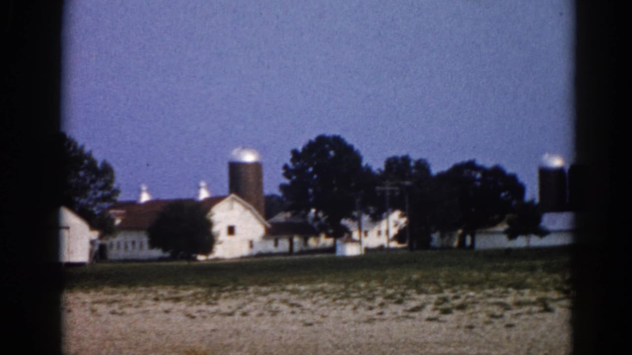 8mm film frame circa 1962 showing an Illinois farm with white outbuildings, prominent grain silos, and mature trees at twilight.