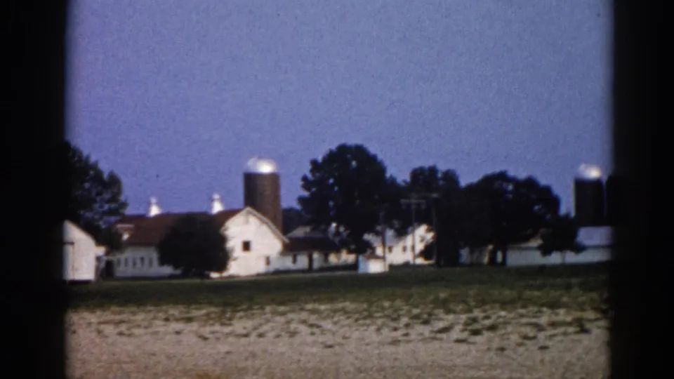 8mm film frame circa 1962 showing an Illinois farm with white outbuildings, prominent grain silos, and mature trees at twilight.