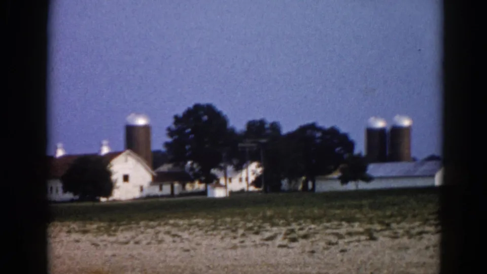 8mm film frame circa 1962, likely Illinois, showing a farm compound with a tall grain silo, white outbuilding, and trees at twilight.