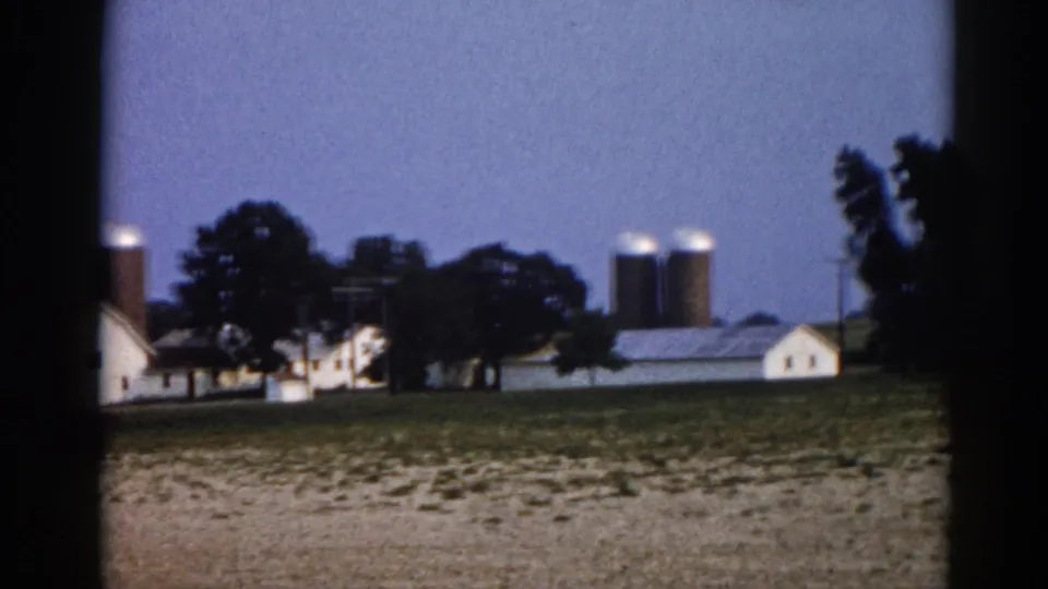 8mm film frame circa 1962 showing an Illinois farm at dusk with paired grain silos, white outbuildings, and trees against a blue evening sky.