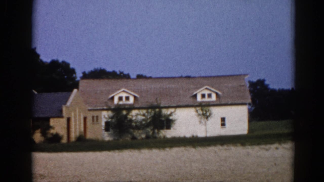 White clapboard farmhouse with two dormer windows at dusk, dry grassy field in foreground, deciduous trees behind.