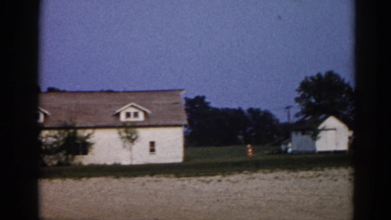 White farmhouse with dormer windows, pan slightly right revealing a second white outbuilding; dusk sky and trees in background.