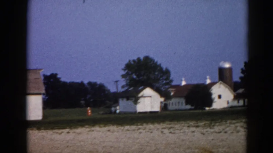 1962 Illinois farmstead wide shot: multiple white outbuildings with red-brown roofs, cylindrical grain silo with bright dome, mature trees, blue dusk sky, amber ground-level lights.