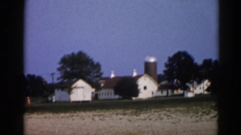 1962 Illinois farmstead pan end: grain silo prominent at right, spread of white outbuildings and barns, blue dusk sky, small amber light near structures.