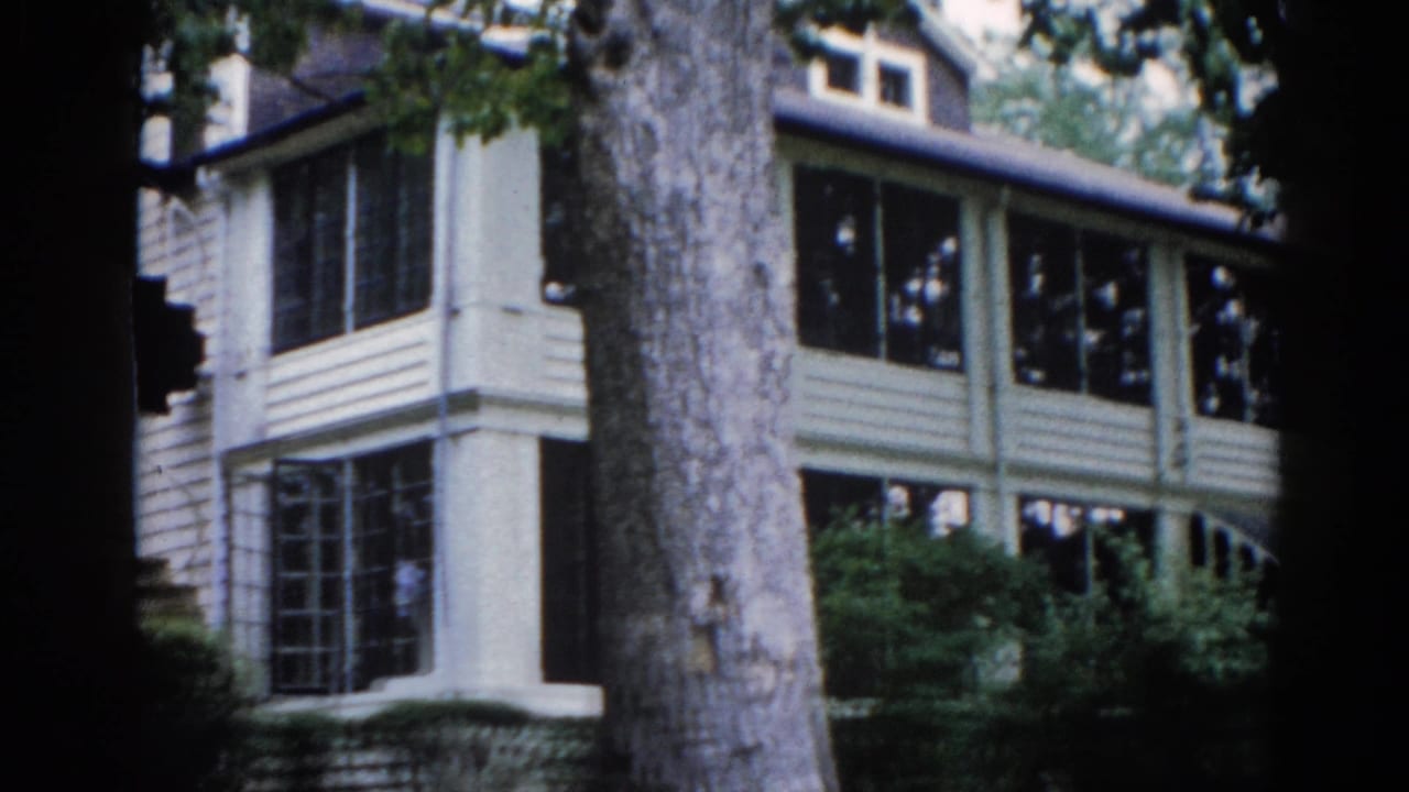 Two-story clapboard house with screened upper windows, a large mature tree trunk in the foreground — 8mm home movie, Illinois, 1962.