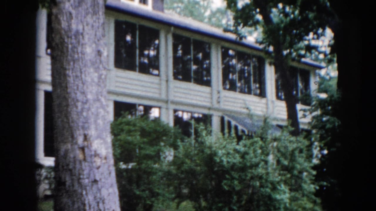 Same two-story clapboard house with large tree trunk in foreground, camera beginning a slow pan across the yard.