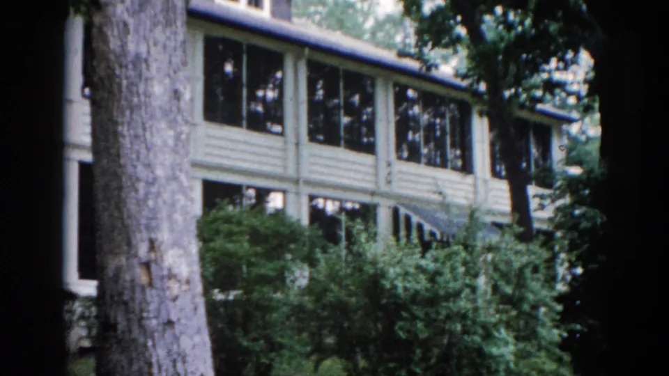 Two-story clapboard house with multi-pane upper windows, large tree trunk prominent in foreground, camera beginning to pan — 8mm home movie, Illinois, 1962.