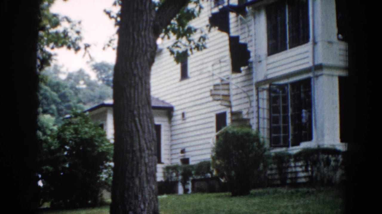 Two-story white clapboard house seen through large tree trunks, screened porch windows on both floors, green lawn below, 8mm film.