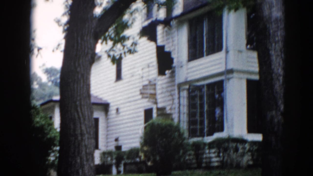 Pan continues across the white-sided Illinois house exterior circa 1962, large tree trunk dominating the foreground, screened porches and clapboard detail visible mid-frame.
