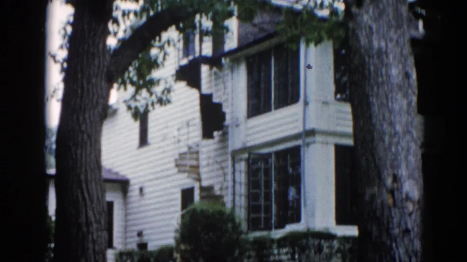 Tighter mid-pan framing of the Illinois house in 1962, tree trunk prominent in foreground, lower screened porch and multi-pane windows visible alongside characteristic 8mm emulsion aging.