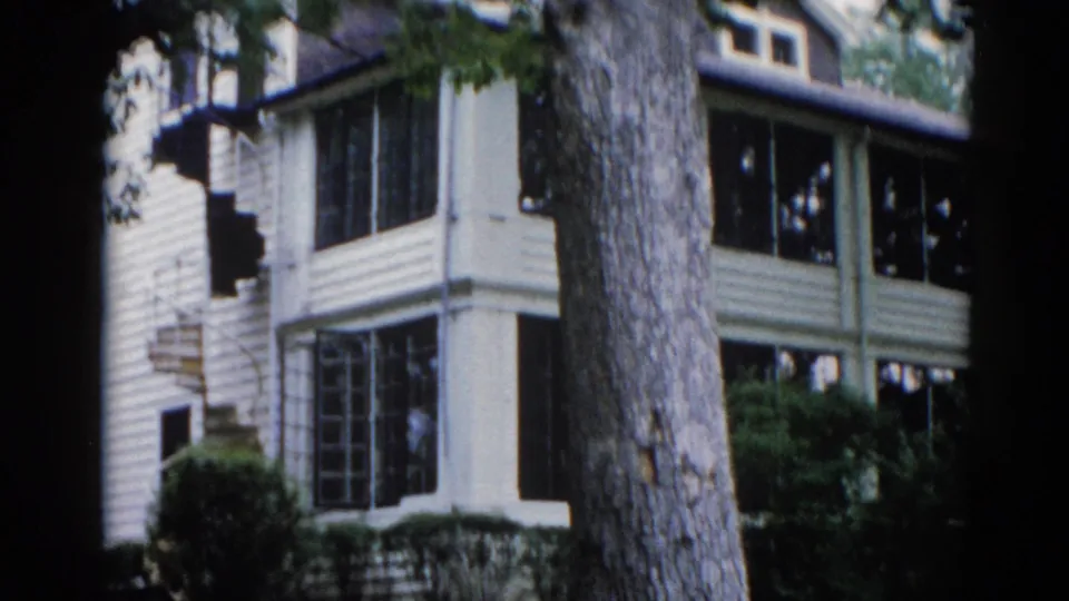 Final frame of a slow pan across an Illinois two-story house in 1962, revealing screened-in porch sections on both floors framed by tree branches and green foliage.