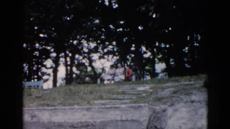 8mm home movie frame from 1962 Illinois showing a small group seated on a grassy hillside near a concrete retaining structure, one figure in a red shirt.