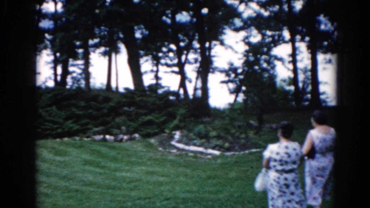 Two women in patterned dresses walking away from camera across a manicured lawn, dense trees and a glimpse of water in the background.