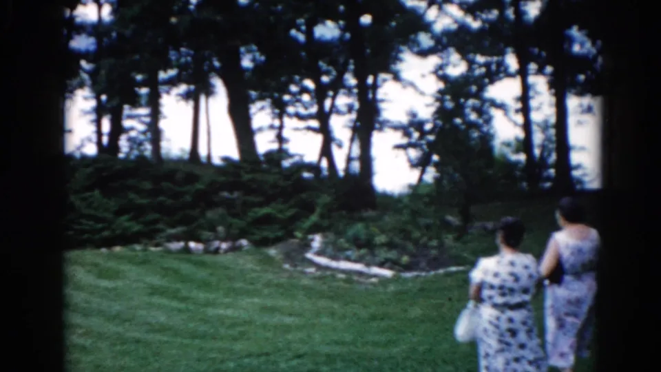 Two women in sleeveless patterned dresses stroll across a manicured garden lawn, shot from behind; recorded as Illinois, circa 1962.