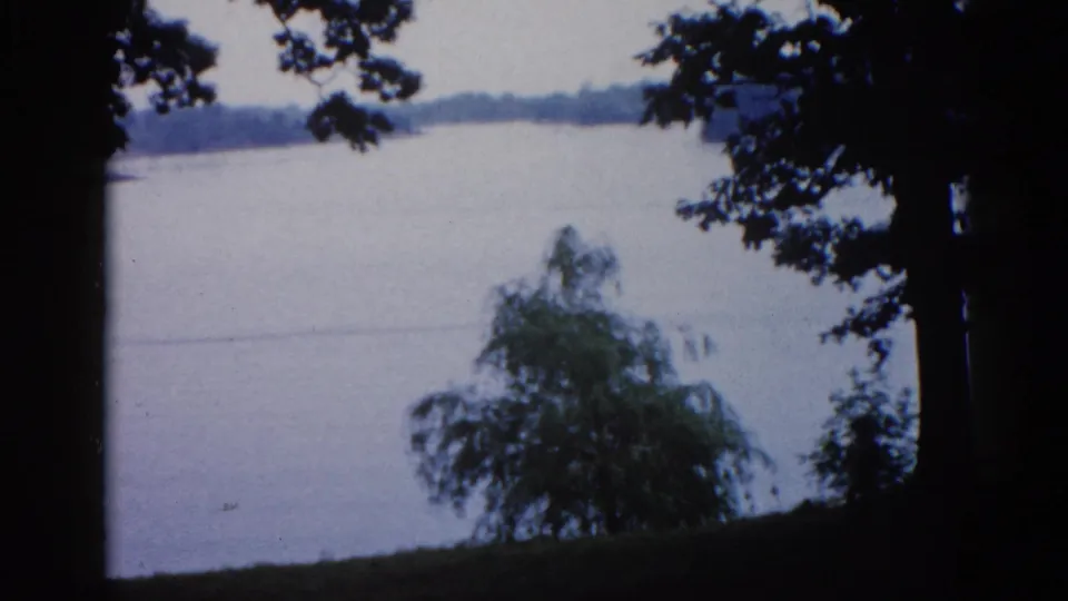 A calm lake framed by overhanging tree branches captured on 8mm film; consistent with an Illinois lakeside setting described as circa 1962.
