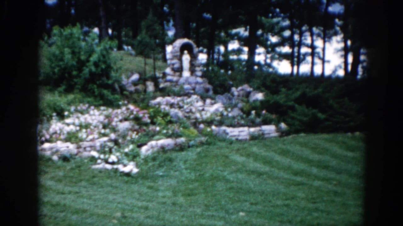 Early 1960s residential garden grotto in Illinois: stone arch shrine, rock-edged flower beds, and mowing-striped lawn on 8mm film.