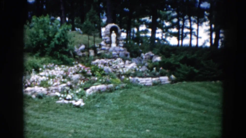 Early 1960s residential garden grotto in Illinois: stone arch shrine, rock-edged flower beds, and mowing-striped lawn on 8mm film.