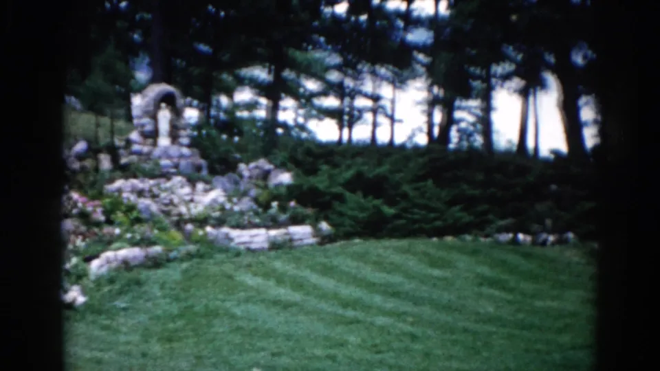 1962 Illinois garden on 8mm: stone grotto at frame edge, striped lawn, dense conifer trees, and a briefly visible figure near the shrine.