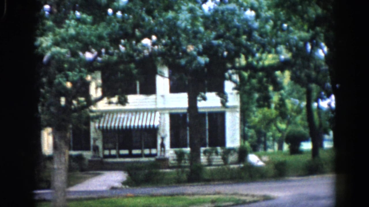 White clapboard house with black-and-white striped awning viewed through mature deciduous trees, 8mm home movie.