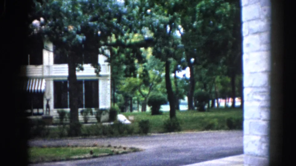 Pan-right view of white clapboard house and striped awning with a stone column visible at the right edge, Illinois, circa 1962 8mm footage.