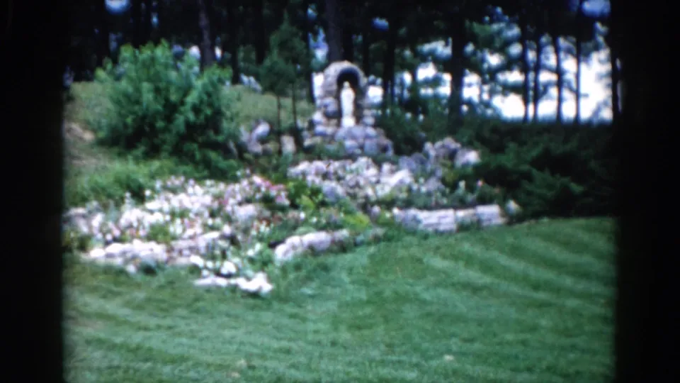 Stone garden grotto with arched niche housing a small statue figure, stone-bordered flower beds, and tall trees behind, Illinois, circa 1962 8mm.