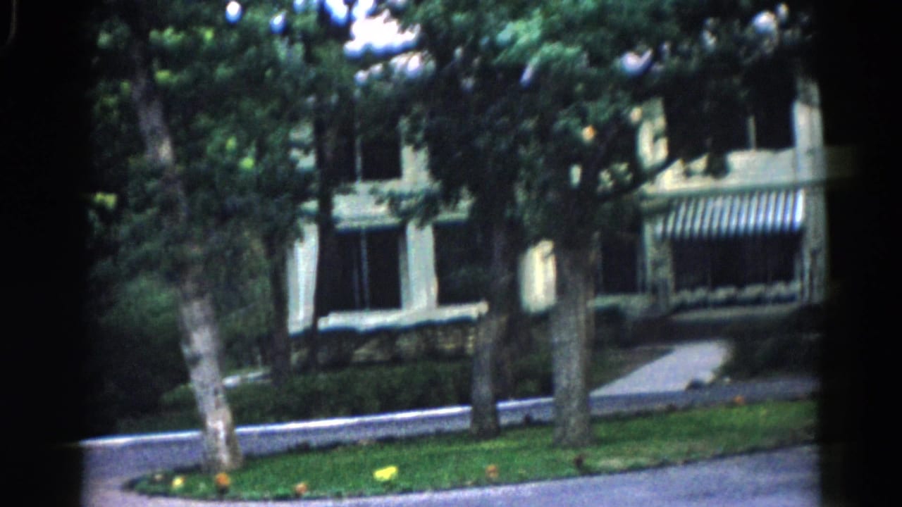 8mm home movie frame, c. 1962 Illinois — white building with black-and-white striped canvas awnings, mature trees, and paved driveway in summer.