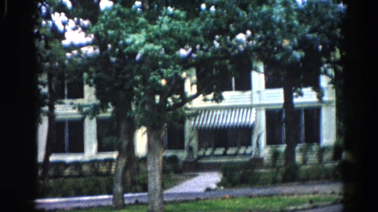 White building with striped awnings partially obscured by mature trees, paved driveway in foreground, static framing similar to opening shot.